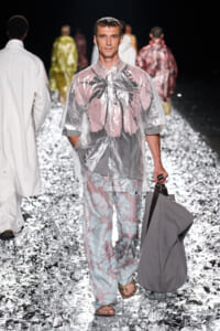 Male model walks the fashion runway in a translucent pink-gray rain poncho with palm print, carrying a gray bag on his right hand.