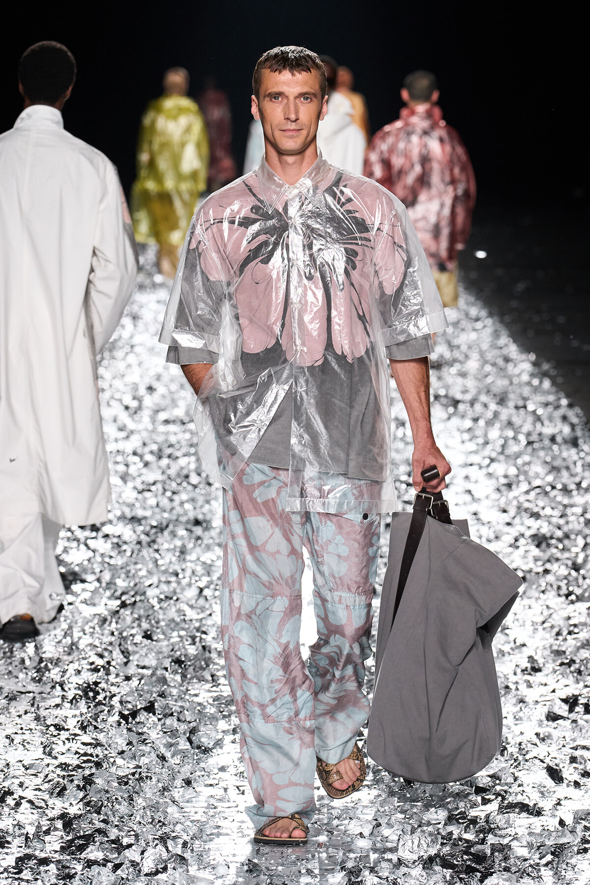 Male model walks the fashion runway in a translucent pink-gray rain poncho with palm print, carrying a gray bag on his right hand.