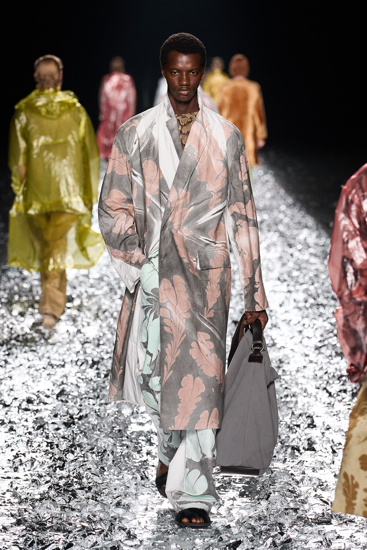 Male runway model in a pale pink and gray floral overcoat, carrying a gray bag on a glittery runway surface.