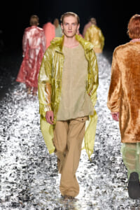 Male model walking toward camera in lime-green metallic coat over sand-toned tunic and khaki cargo pants on a reflective silver runway.
