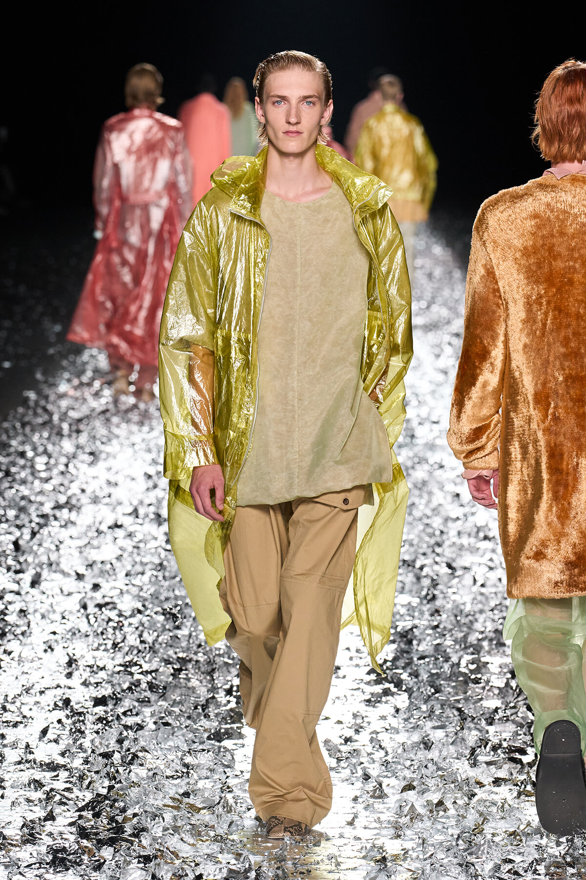 Male model walking toward camera in lime-green metallic coat over sand-toned tunic and khaki cargo pants on a reflective silver runway.