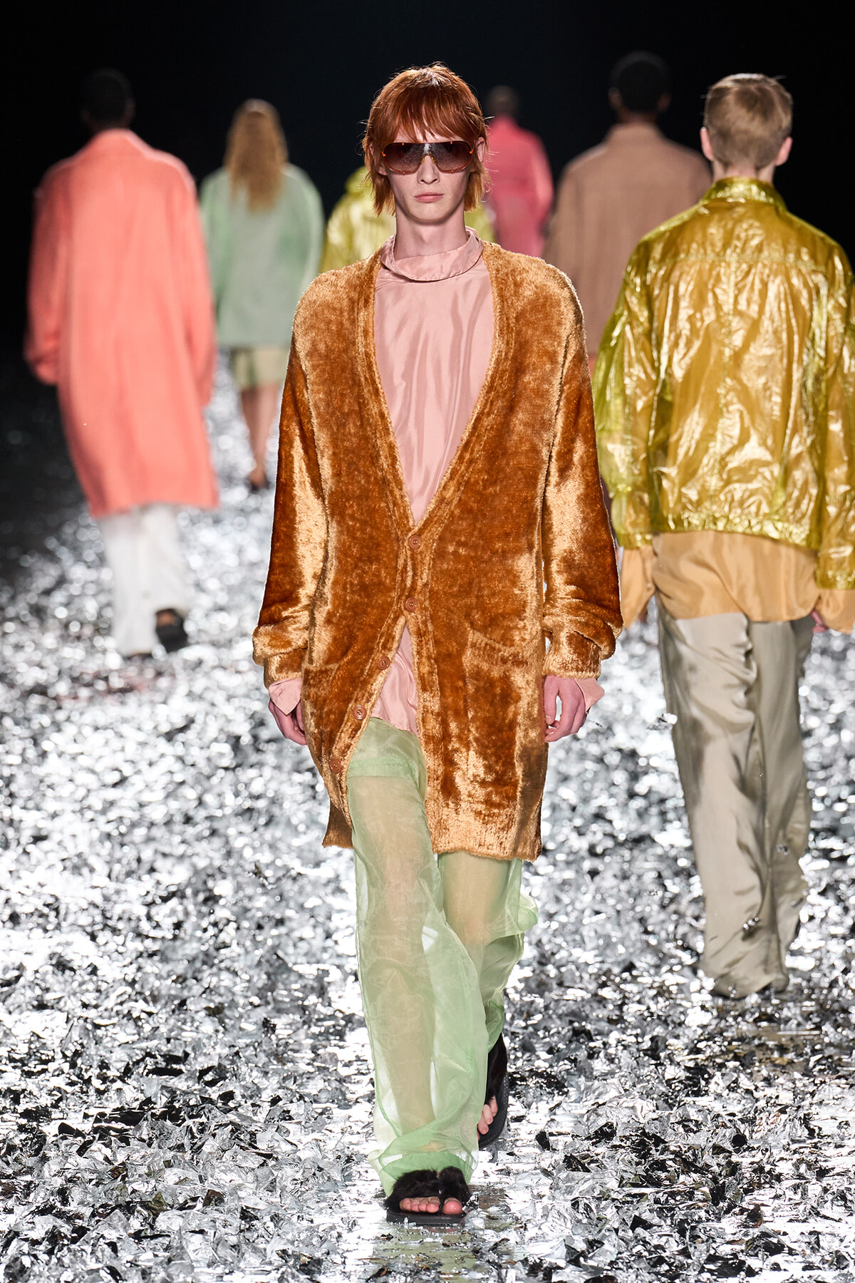 Model on a runway wearing a fuzzy orange cardigan over a pink top and pale green pants, sunglasses, walking through metallic confetti.