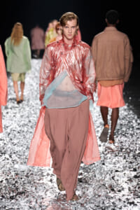 Model walks a runway in a pink metallic trench over a sheer blue layer and dusty-rose pants, with silver confetti on the floor.
