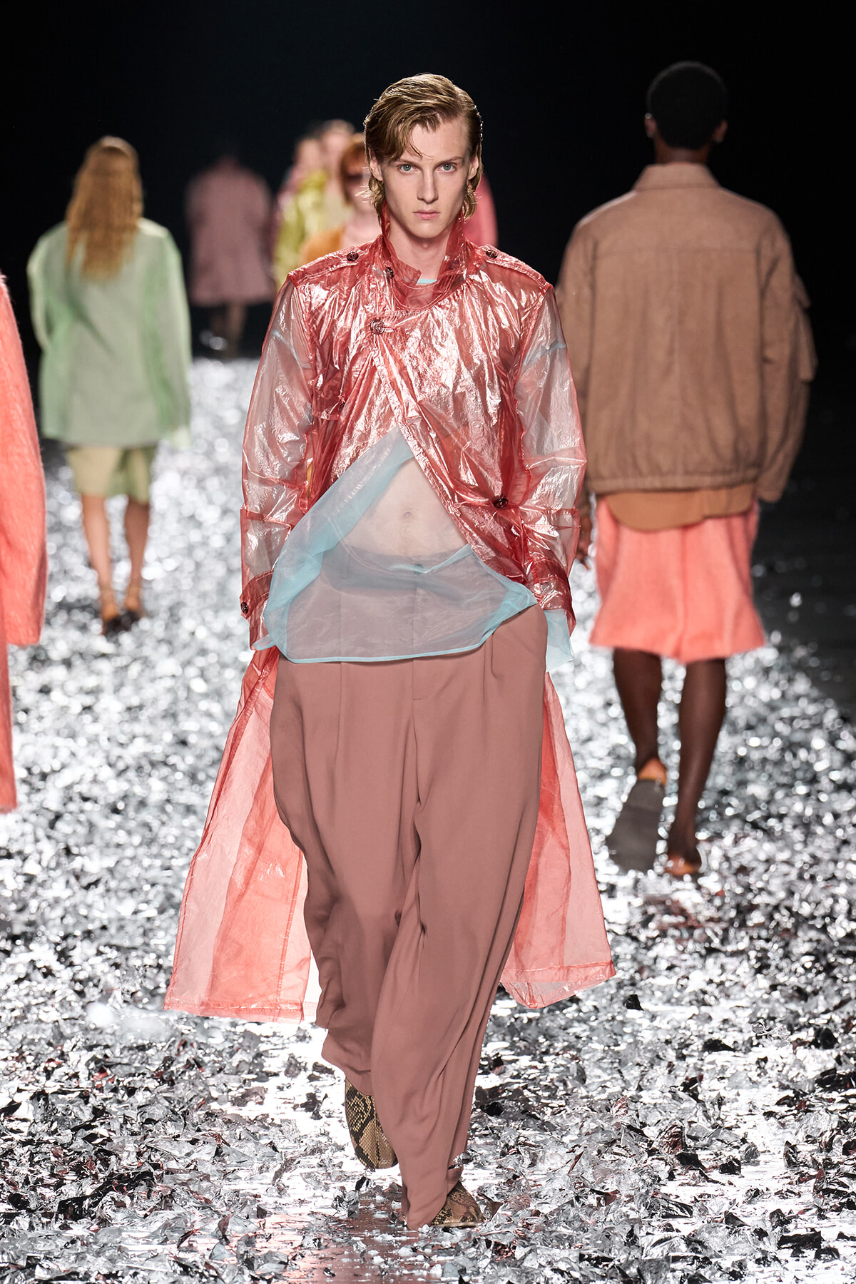 Model walks a runway in a pink metallic trench over a sheer blue layer and dusty-rose pants, with silver confetti on the floor.