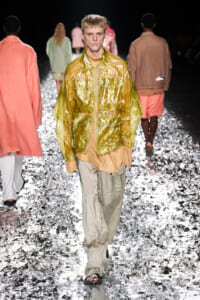 Model walks the runway in a metallic gold translucent jacket over a beige top and pale pants, silver confetti on the floor around him.