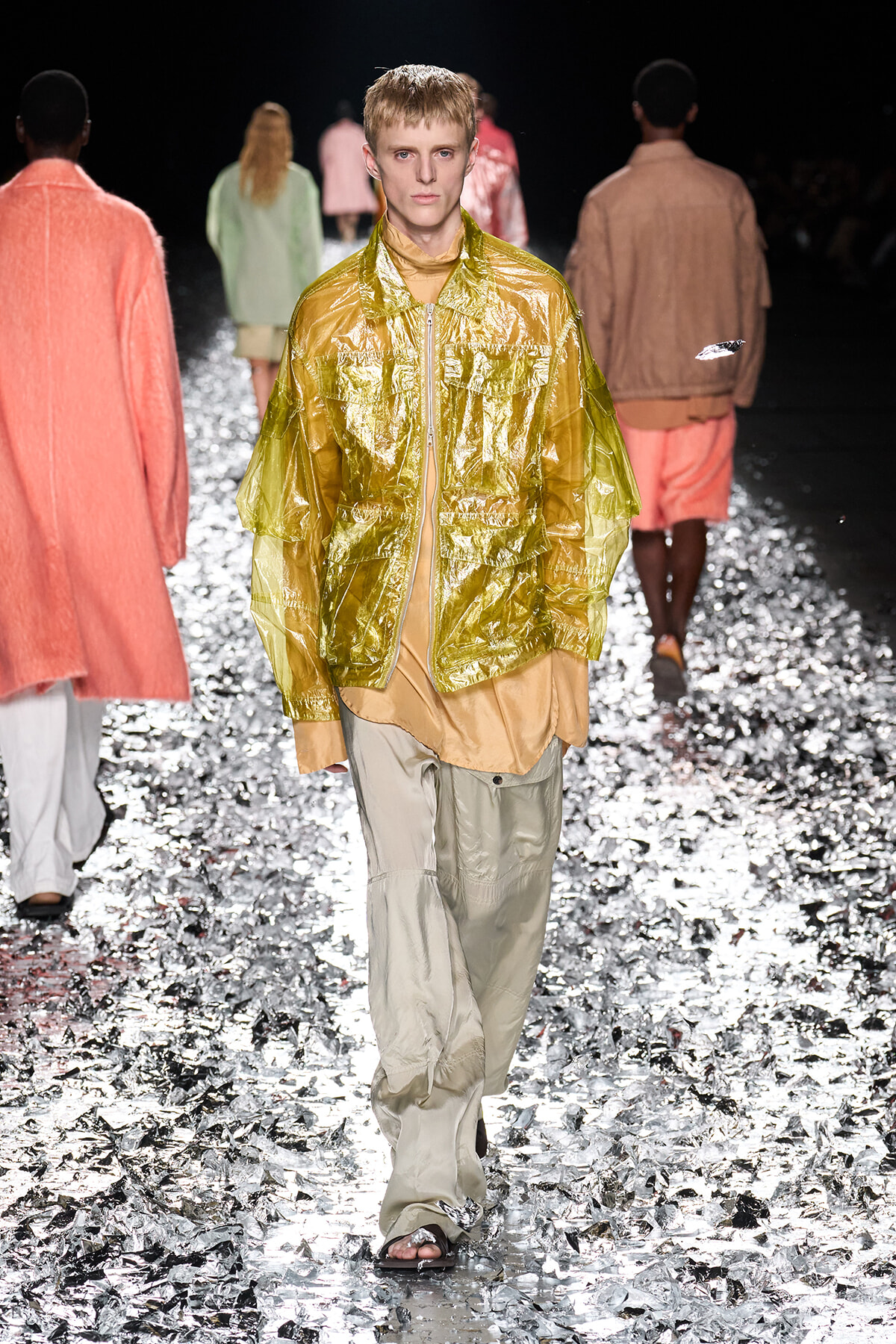 Model walks the runway in a metallic gold translucent jacket over a beige top and pale pants, silver confetti on the floor around him.