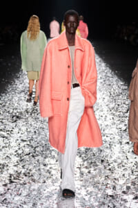 Model walking a runway in a oversized pink fuzzy coat, light top, and white trousers; silver confetti on the floor adds a festive backdrop.