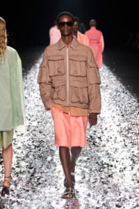 Model on a fashion runway wearing a tan utility jacket with numerous pockets, pink shorts, and dark sunglasses on a glittering catwalk surface.
