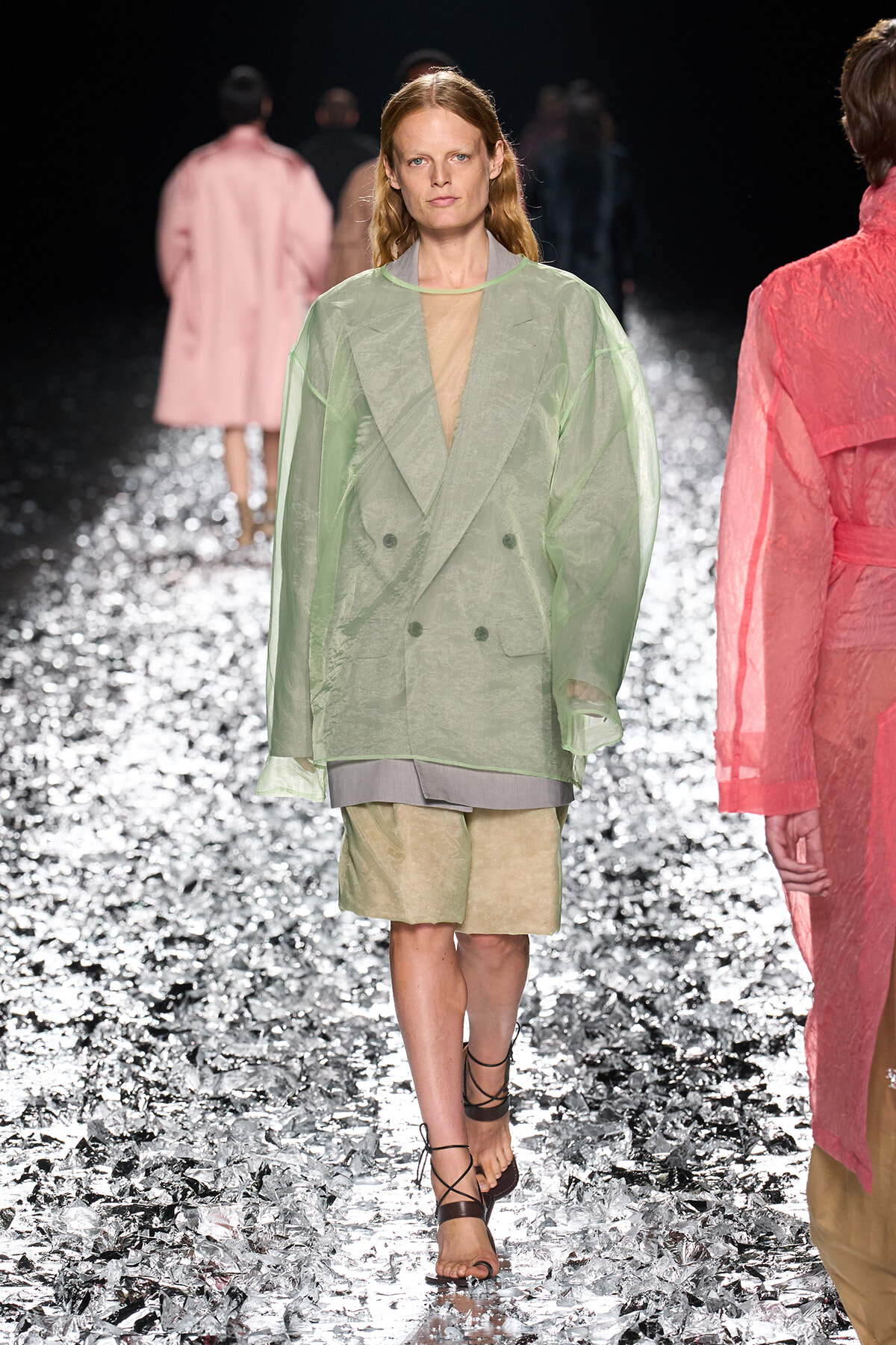 Model walks a fashion runway in a sheer, oversized light-green blazer over layered beige shorts, glittering stage behind.