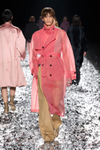 Model walks a pink sheer trench coat belted at the waist over tan wide-leg pants on a glittery runway surface.