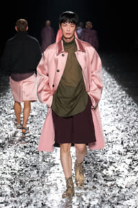 Model walking the runway in a pink satin long coat over an olive green top and burgundy shorts, paired with snakeskin boots, on a glittery runway surface.
