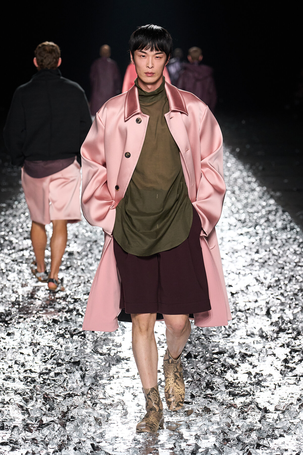 Model walking the runway in a pink satin long coat over an olive green top and burgundy shorts, paired with snakeskin boots, on a glittery runway surface.