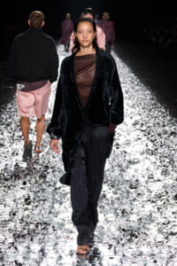 Model walks down a glittering runway wearing a black faux-fur cardigan over a sheer brown top and black cargo pants on a reflective, foil-strewn catwalk.