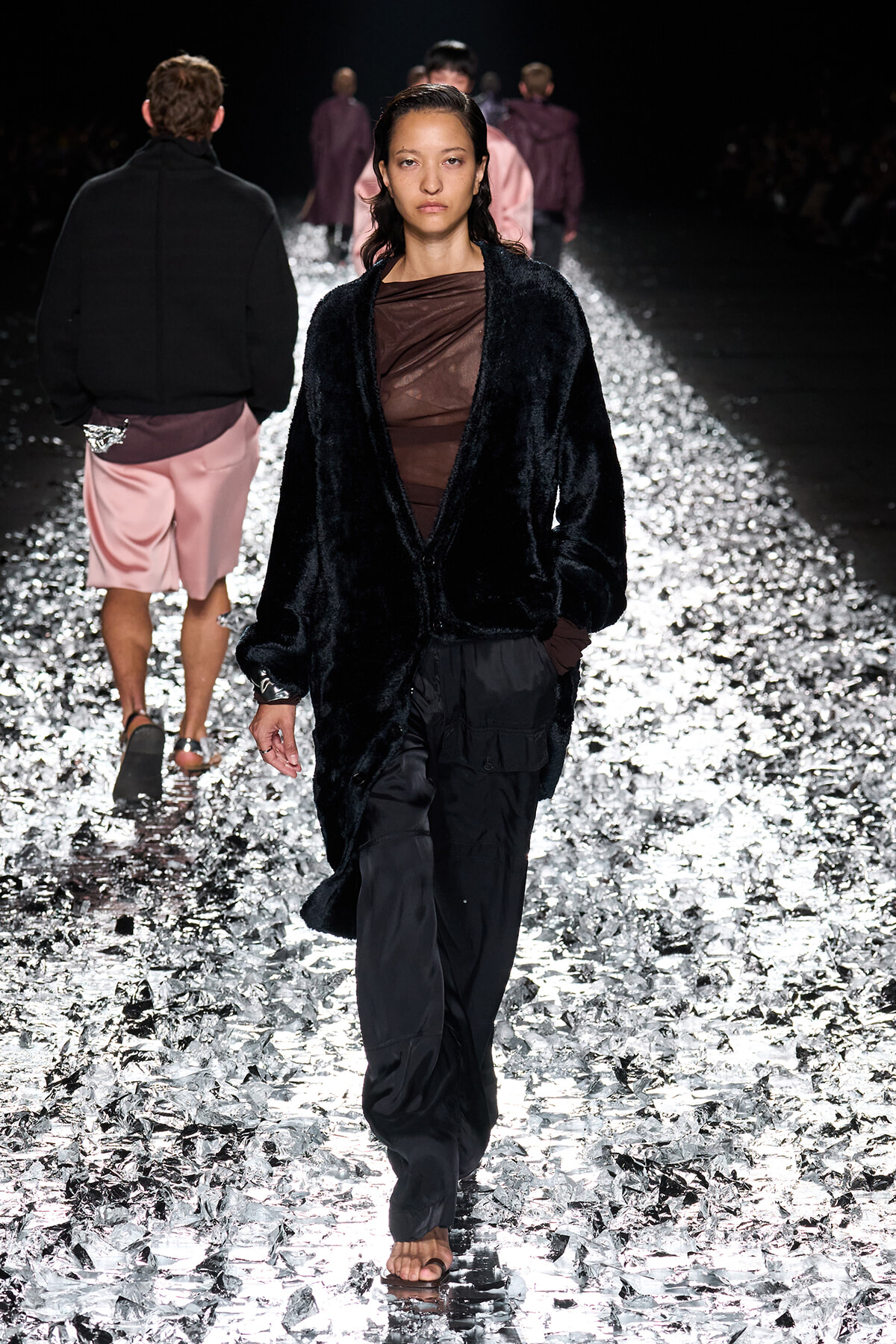 Model walks down a glittering runway wearing a black faux-fur cardigan over a sheer brown top and black cargo pants on a reflective, foil-strewn catwalk.