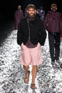 Male model walking a fashion-show runway in a black zippered sweater, pink shorts, and brown sandals on a metallic-speckled floor.