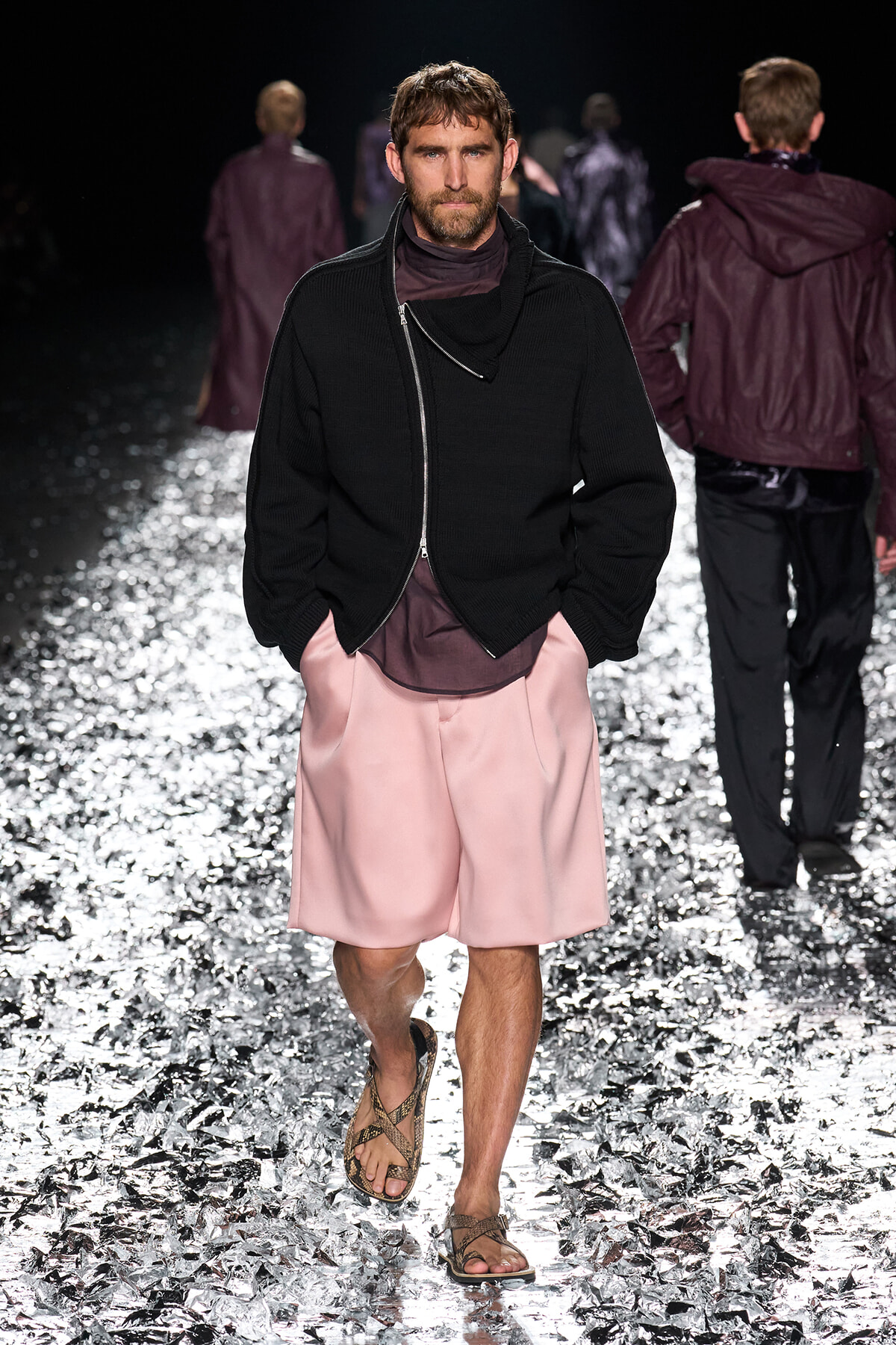 Male model walking a fashion-show runway in a black zippered sweater, pink shorts, and brown sandals on a metallic-speckled floor.
