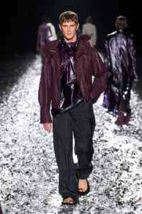 Male model on a glittering runway wearing a maroon hooded jacket over a shiny purple top and black cargo pants, barefoot.
