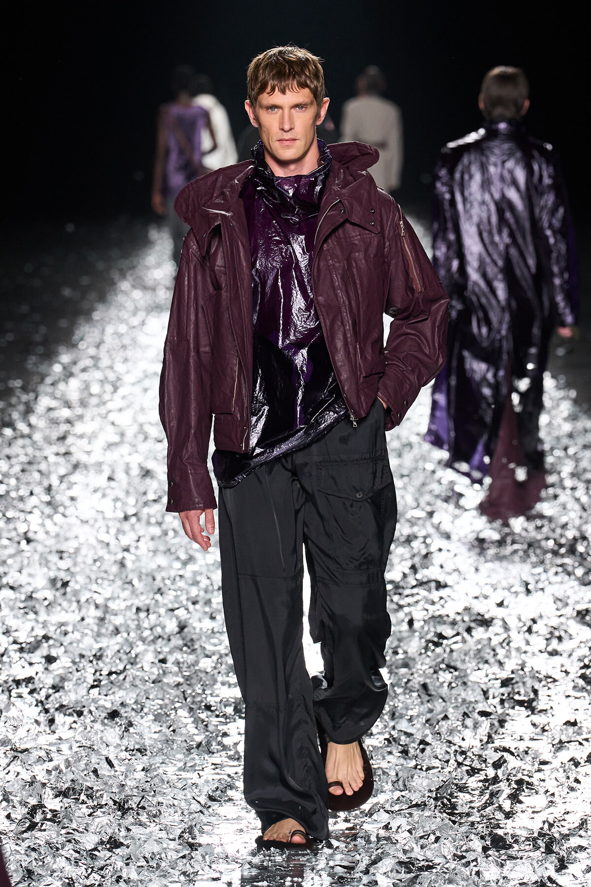Male model on a glittering runway wearing a maroon hooded jacket over a shiny purple top and black cargo pants, barefoot.