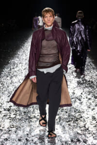 Model walking a fashion show runway in a burgundy long coat over a sheer brown top and dark pants, with sandals, on a glittering reflective floor.