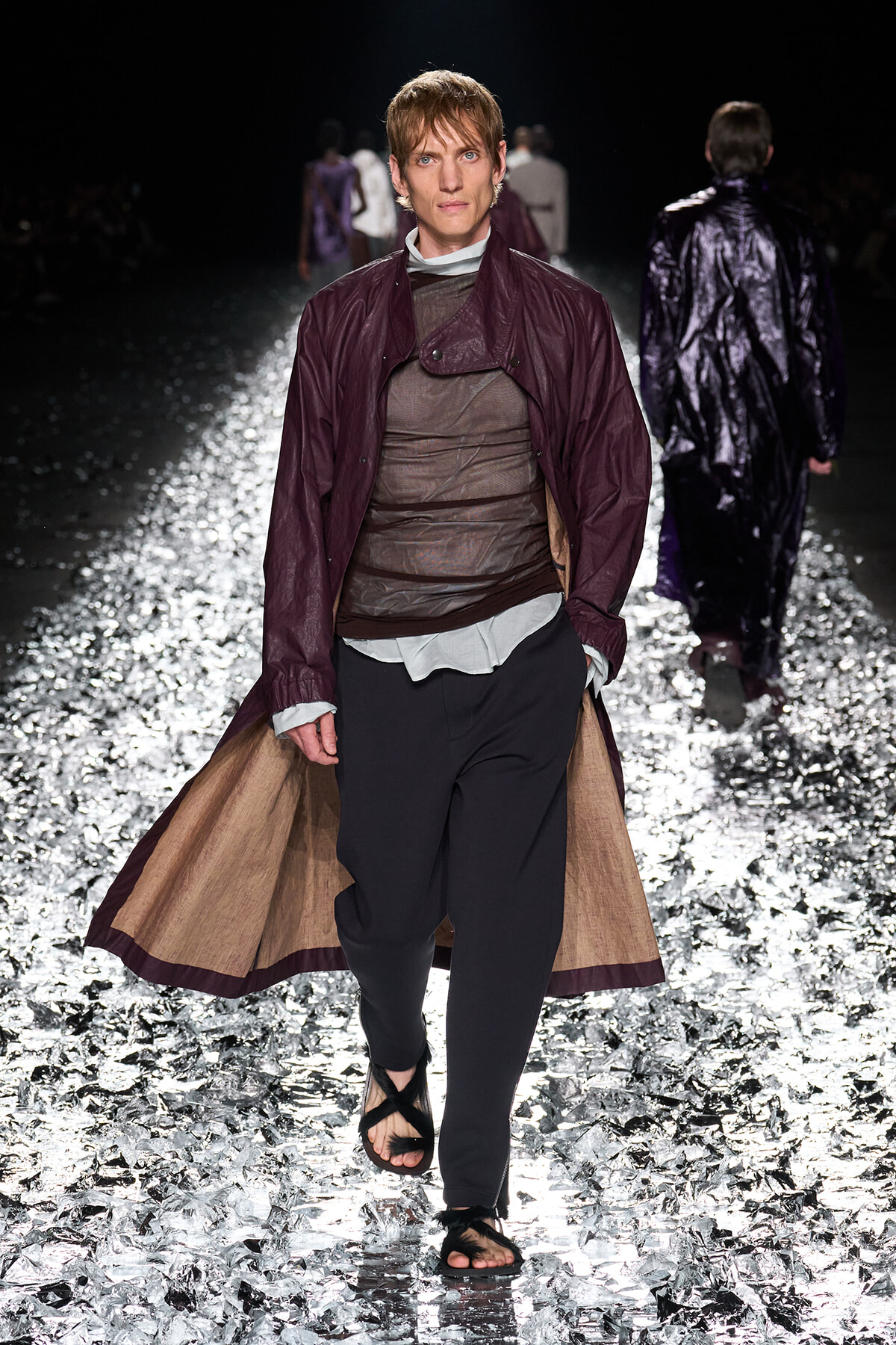 Model walking a fashion show runway in a burgundy long coat over a sheer brown top and dark pants, with sandals, on a glittering reflective floor.