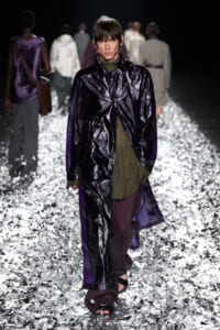 Model walking a fashion-show runway in a glossy dark purple coat over an olive-green top and burgundy pants, on a reflective metallic-foil floor.