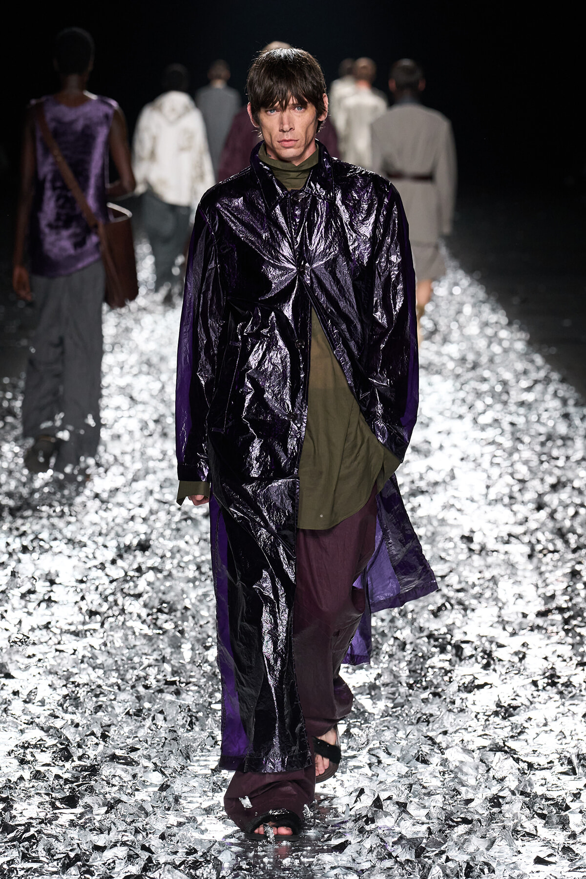 Model walking a fashion-show runway in a glossy dark purple coat over an olive-green top and burgundy pants, on a reflective metallic-foil floor.