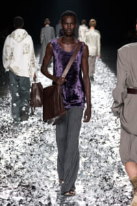 Male fashion model on a runway wearing a purple velvet sleeveless top, gray wide-leg pants, and a brown crossbody bag.