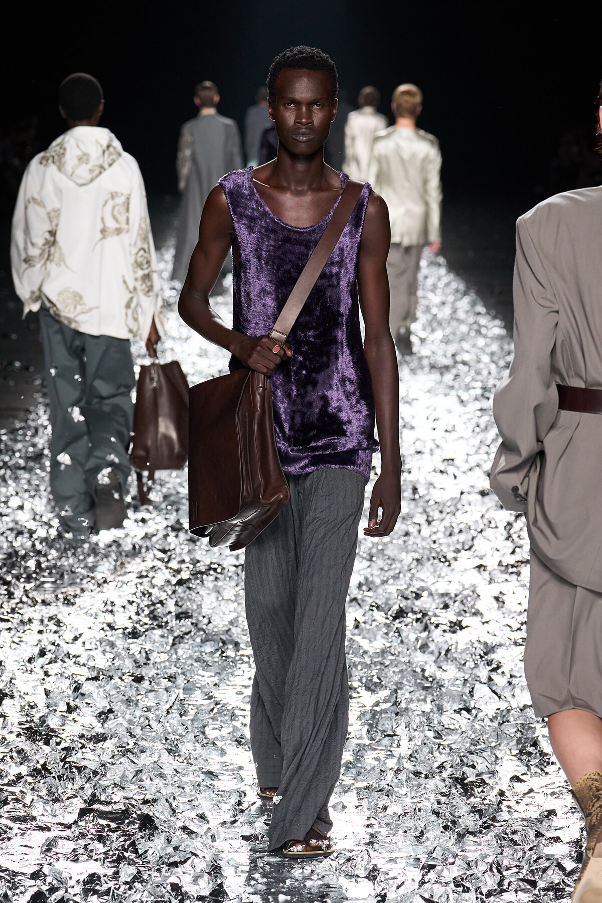 Male fashion model on a runway wearing a purple velvet sleeveless top, gray wide-leg pants, and a brown crossbody bag.