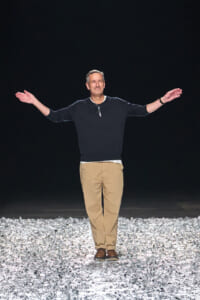 Man stands on a glittering rocky runway at night with arms outstretched, wearing a dark long-sleeve shirt and tan pants.