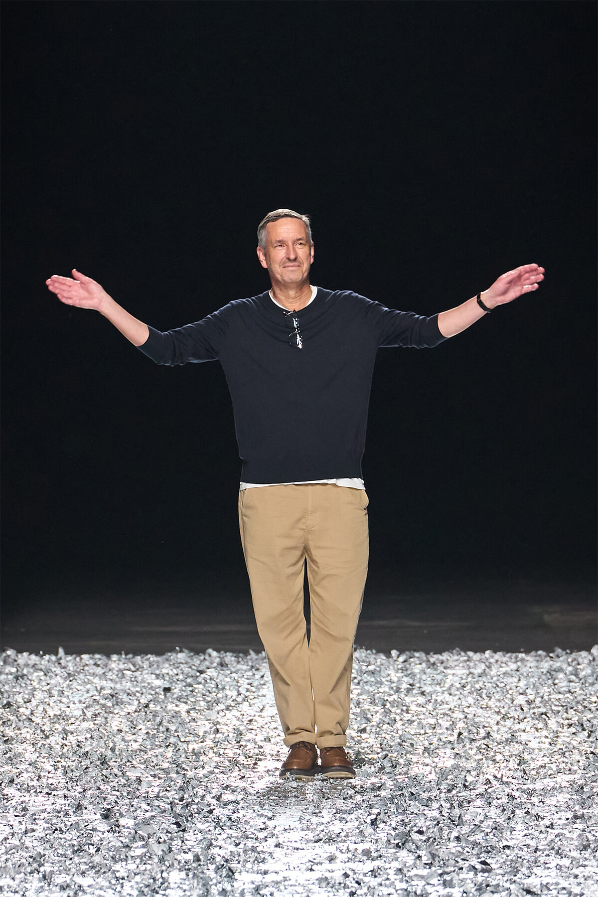 Man stands on a glittering rocky runway at night with arms outstretched, wearing a dark long-sleeve shirt and tan pants.