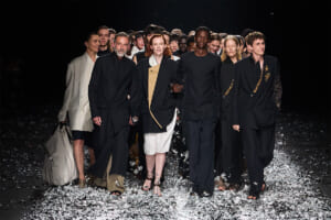 Group of diverse models walking arm in arm on a fashion runway, wearing black and neutral-toned outfits with metallic confetti on the floor behind them.