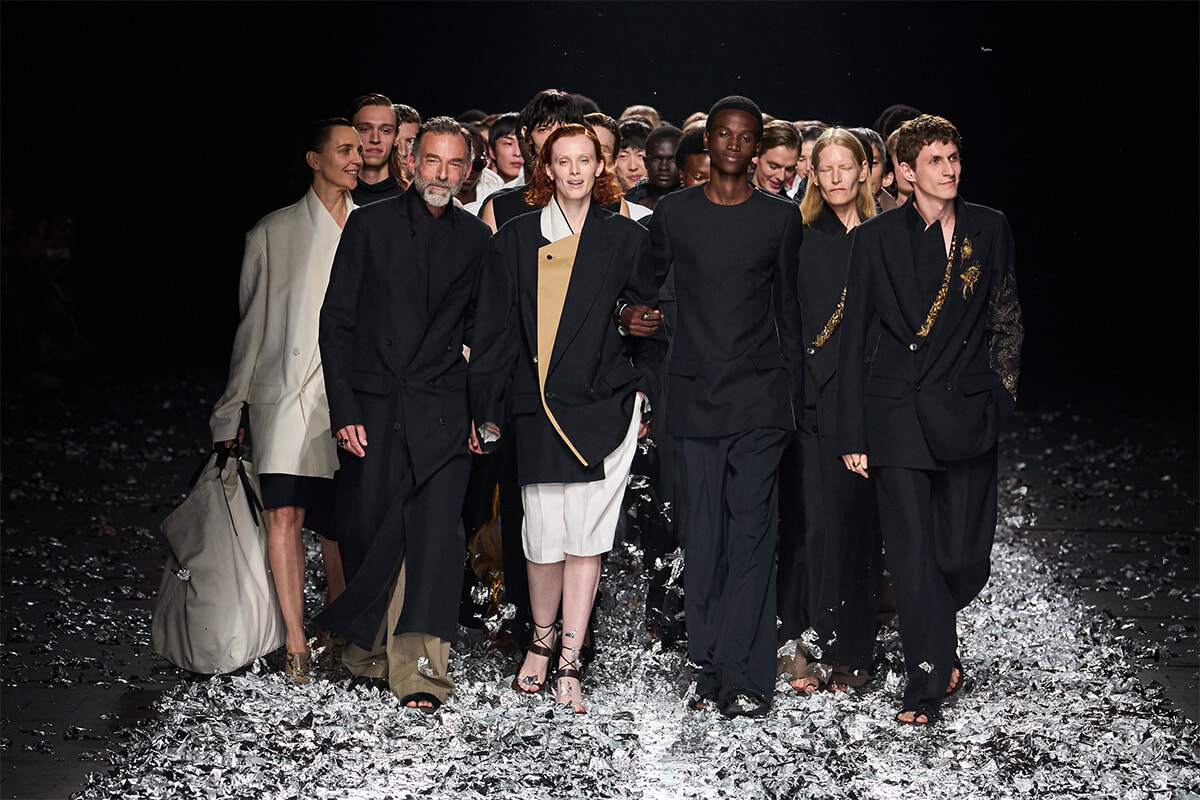 Group of diverse models walking arm in arm on a fashion runway, wearing black and neutral-toned outfits with metallic confetti on the floor behind them.