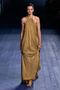Model walking a runway in a sleeveless, gold metallic halter-neck gown with gathered draping and matching sandals.