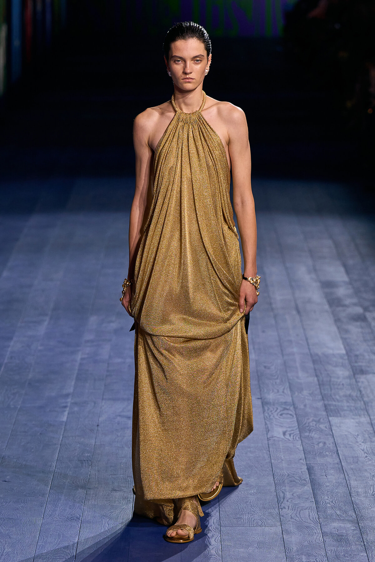 Model walking a runway in a sleeveless, gold metallic halter-neck gown with gathered draping and matching sandals.