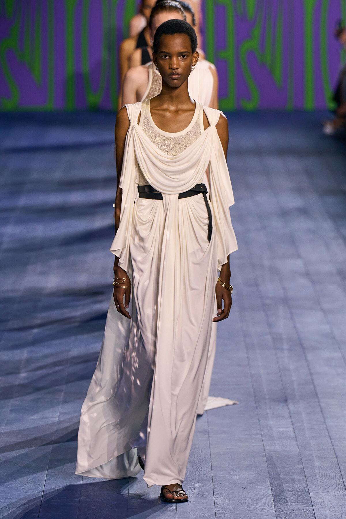 Model walks the runway in a cream draped gown with a black belt and gold jewelry.