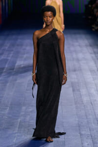 Model walks the runway in a shimmering black one-shoulder gown with a draped scarf and statement bracelets.