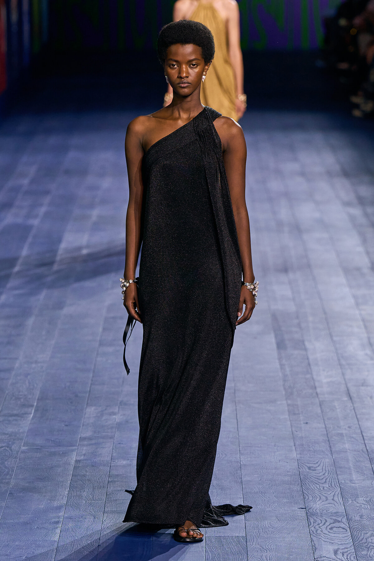 Model walks the runway in a shimmering black one-shoulder gown with a draped scarf and statement bracelets.