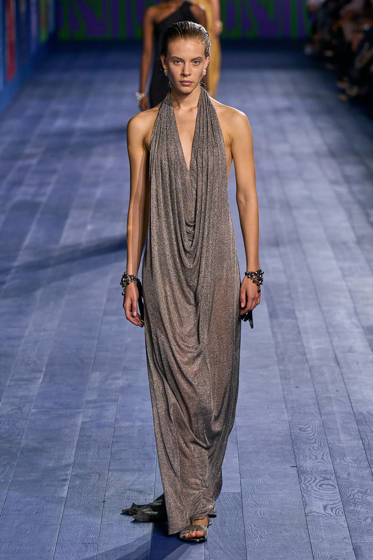 Model walking a fashion runway in a shimmering metallic draped halter gown with a deep V neckline, slicked-back hair, and crystal bracelets.