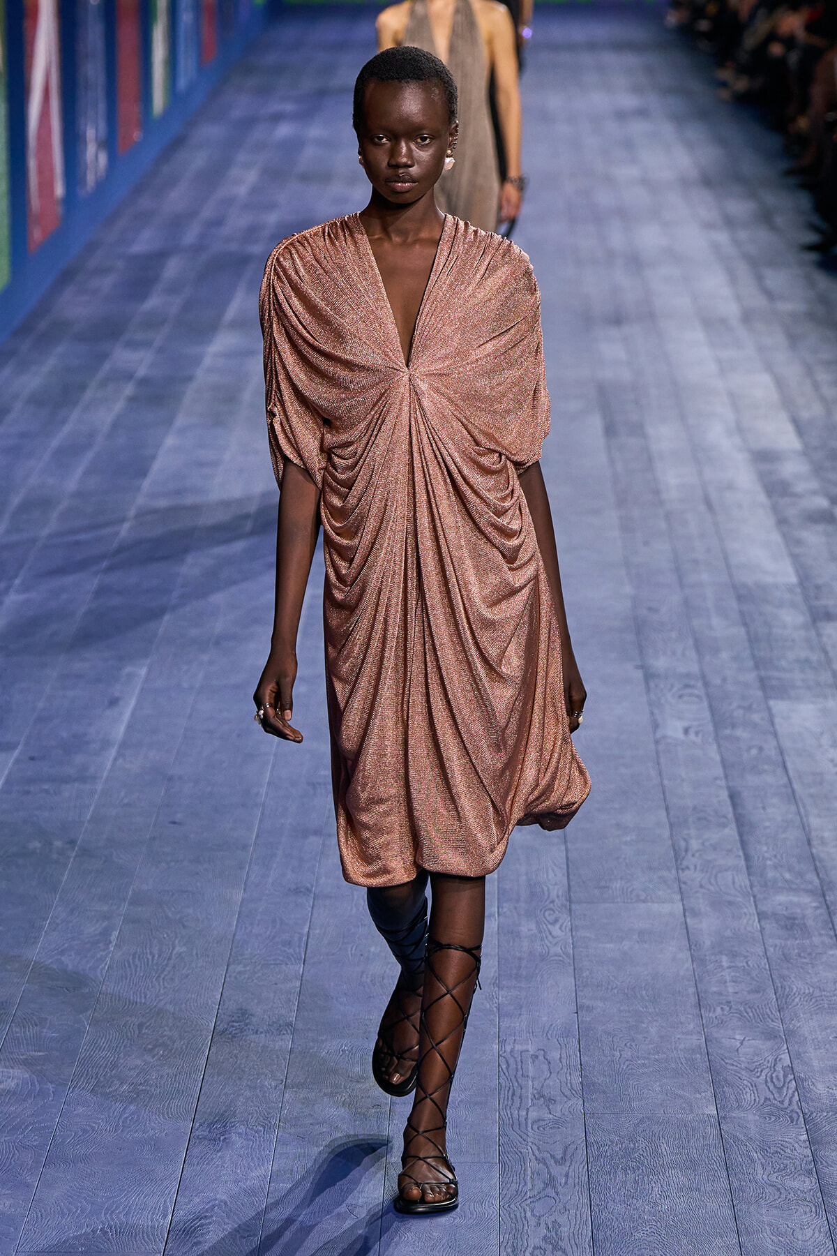 Female model on a runway wearing a shimmery rose-gold draped knee-length dress with a central knot and gladiator-style lace-up sandals.