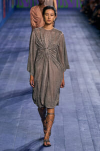 Model walking runway in a metallic taupe draped dress with a twisted knot at the waist and gladiator sandals.