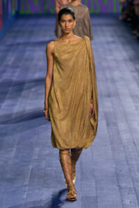Model walks the runway in a shimmery gold draped one-shoulder dress with a bubble hem and strappy sandals.