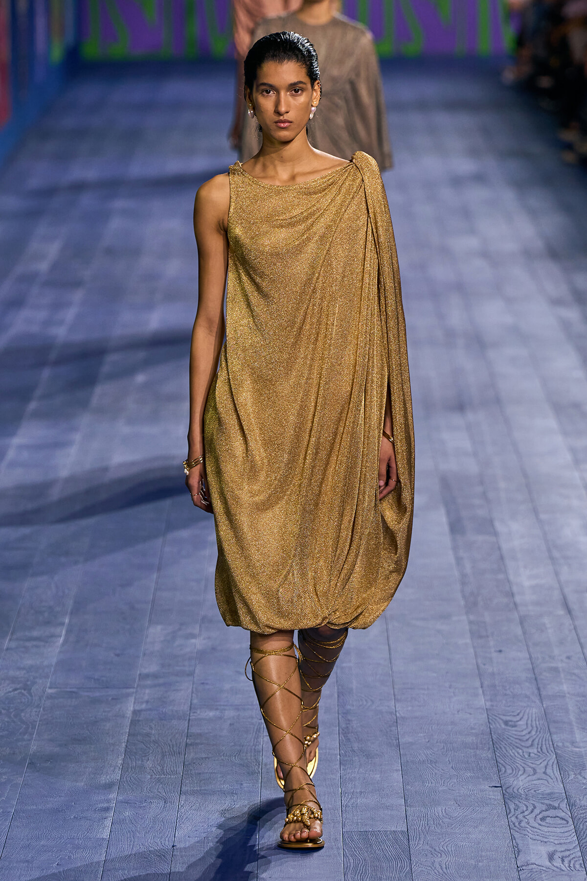 Model walks the runway in a shimmery gold draped one-shoulder dress with a bubble hem and strappy sandals.