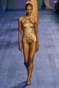 Runway model wearing a metallic gold, scale-textured halter bodysuit with lace-up gladiator sandals walking down a blue fashion show runway.