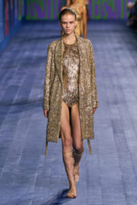 Model walks a fashion runway wearing a full-length gold sequin coat over a metallic, embellished dress and gold gladiator sandals.
