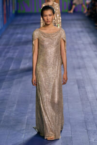 Model walks a glittering gold gown with draped neckline on a fashion runway.