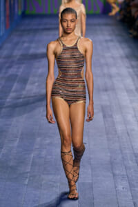 Female model walking a fashion runway in a sleeveless metallic striped bodysuit with side cutouts and lace-up sandals.