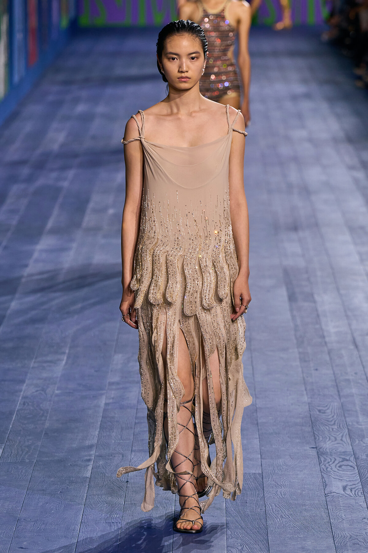 Model walking a runway in a nude-beige strapless gown with cascade-sequin fringe and gladiator-style sandals.