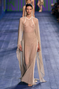 Model walks the runway in a sheer, nude-toned gown with a matching cape, light sparkly fabric and open sides at the ankles