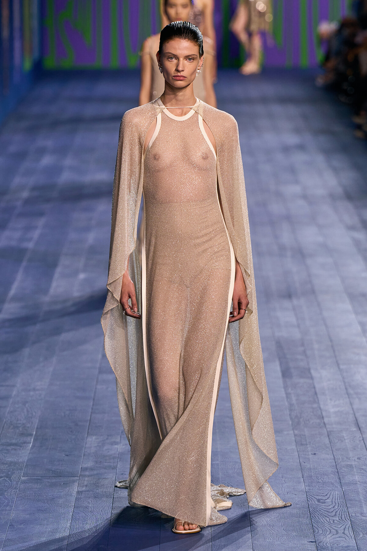 Model walks the runway in a sheer, nude-toned gown with a matching cape, light sparkly fabric and open sides at the ankles
