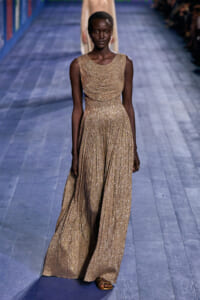 Model walking runway in a gold, glittery sleeveless gown with a pleated draped bodice and full skirt, wearing flat sandals.